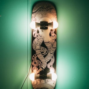 Op de afbeelding: Een skateboard wandlamp met een zwart-wit octopus ontwerp. Het skateboard heeft twee lampen met elk twee lampen. De lamp is bevestigd aan een turquoise muur.