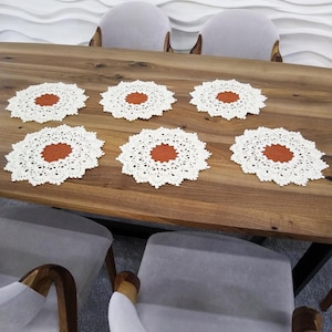 Könnte beinhalten: Sechs weiße und rostfarbene Häkeluntersetzer auf einem Holztisch angeordnet. Die Untersetzer haben ein kreisförmiges Design mit einem rostfarbenen Zentrum und einem detaillierten, durchbrochenen Rand. Der Tisch ist von grauen Stühlen umgeben.