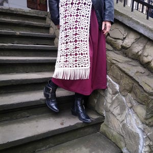 May include: A burgundy skirt with a white, open-weave crochet overlay featuring a floral pattern and fringe. The person is wearing black boots with buckles and a black leather jacket. The image is taken outdoors on stone steps.