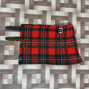 Puede incluir: Un kilt de tartán rojo, verde y blanco con correas de cuero negro y una hebilla plateada. El kilt está doblado y sobre una superficie de madera.