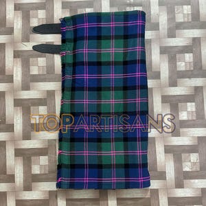 Könnte beinhalten: Ein gefalteter Kilt mit Tartanmuster in Blau, Grün, Schwarz und Pink. Er hat zwei schwarze Lederriemen und einen Fransenrand. Der Text "TOPARTISANS" ist sichtbar. Traditionelles Kleidungsstück.