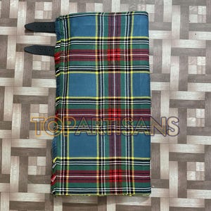 Puede incluir: Un kilt plegado con un patrón de tartán azul, verde, amarillo y rojo. El kilt tiene correas de cuero negro y el texto "TOPARTISANS" es visible. La tela tiene un borde con flecos.