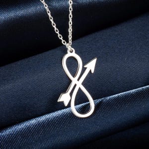 May include: Silver necklace with an infinity symbol design crossed by an arrow. The pendant hangs from a fine chain, set against a dark blue fabric. The arrow points upwards, adding a directional element to the design.