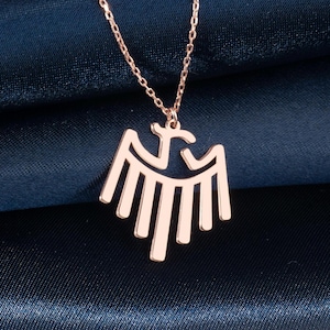 May include: Rose gold necklace featuring a stylized eagle pendant. The pendant has a symmetrical design with a central head and outstretched wings. The chain is delicate, and the necklace is displayed on a dark blue fabric.