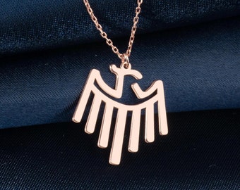 May include: Rose gold necklace featuring a stylized eagle pendant. The pendant has a symmetrical design with a central head and outstretched wings. The chain is delicate, and the necklace is displayed on a dark blue fabric.