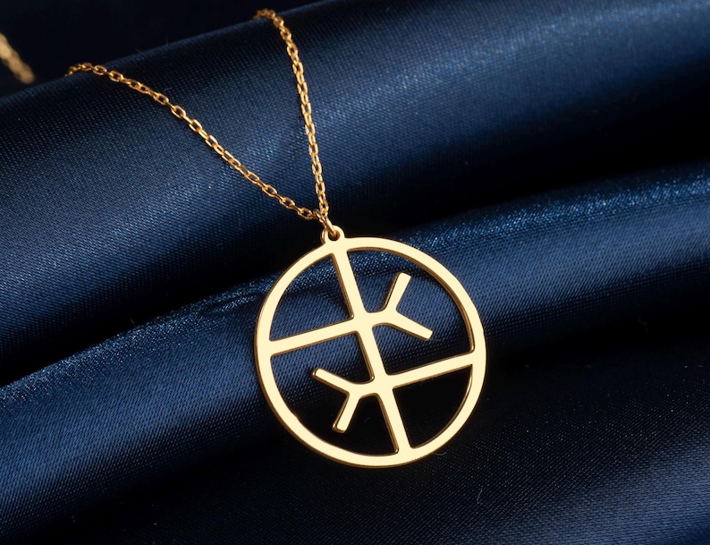 May include: Gold necklace with a circular pendant featuring a geometric design. The pendant is composed of intersecting lines forming a stylized symbol within a circular frame. The chain is delicate and gold-toned. The necklace is displayed against a dark blue fabric backdrop.