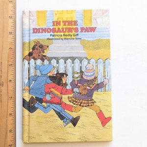 May include: A children's book cover with a yellow background and a blue house. The title is "In the Dinosaur's Paw" by Patricia Reilly Giff, illustrated by Blanche Sims. Three children in winter clothing are running away from a brown and white dog.