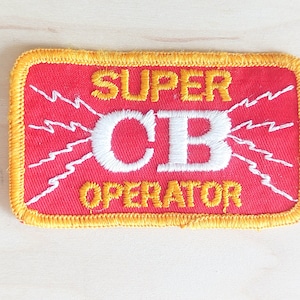 May include: A red and yellow embroidered patch with the text "SUPER CB OPERATOR" in white lettering. The patch features white lightning bolts around the text.
