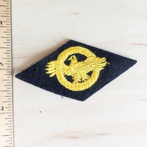 May include: A black felt diamond-shaped patch with a gold embroidered eagle inside a circle.