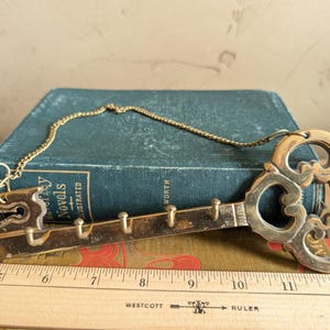 May include: A brass key-shaped wall hook with a decorative design. The key has multiple small hooks and a chain for hanging. The key is approximately 12 inches long.