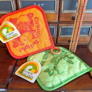 May include: Two quilted pot holders from The Rainbow Collection. One is orange with an orange design and the words "Orange Delight." The other is green with a lime design. Both have wooden ring handles.
