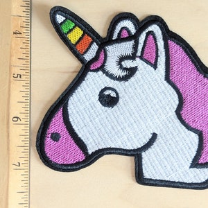 May include: Embroidered iron-on patch of a white unicorn with a rainbow horn and pink mane. The unicorn is facing right and has a black outline.