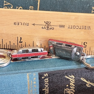 May include: Two miniature harmonica keyrings. The keyrings are silver-toned with red accents and a small metal tag that reads "JAPAN". The keyrings are resting on a stack of books and a wooden ruler.