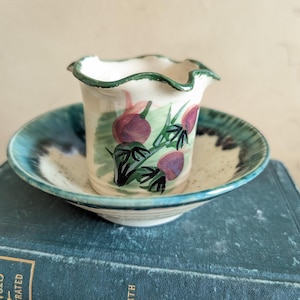 May include: A small, hand-painted ceramic vase with a frilled rim, featuring a design of purple fruit and green leaves. It sits on a matching saucer with a blue and white speckled pattern. The set is displayed on a vintage blue book.