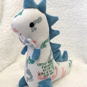 May include: A plush dinosaur toy in white, blue, and pink. The dinosaur has blue spikes, feet, and accents. The toy is personalized with the name "Vittori Ferrari!!!", date, weight, and time of birth.