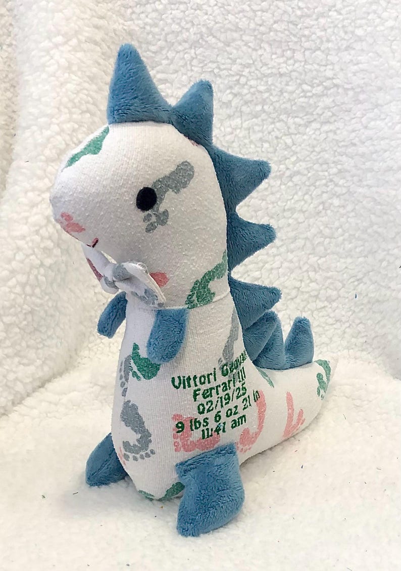 May include: A plush dinosaur toy in white, blue, and pink. The dinosaur has blue spikes, feet, and accents. The toy is personalized with the name "Vittori Ferrari!!!", date, weight, and time of birth.