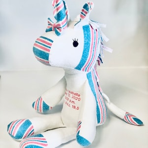 May include: A white unicorn stuffed animal with pink and blue striped fabric. The unicorn has a pink bow on its horn and a black eye. The unicorn is sitting on a white surface. The text "Isabella May 12, 2020 8:08 p.m. 7 oz. 19.5" is printed on the unicorn's body.