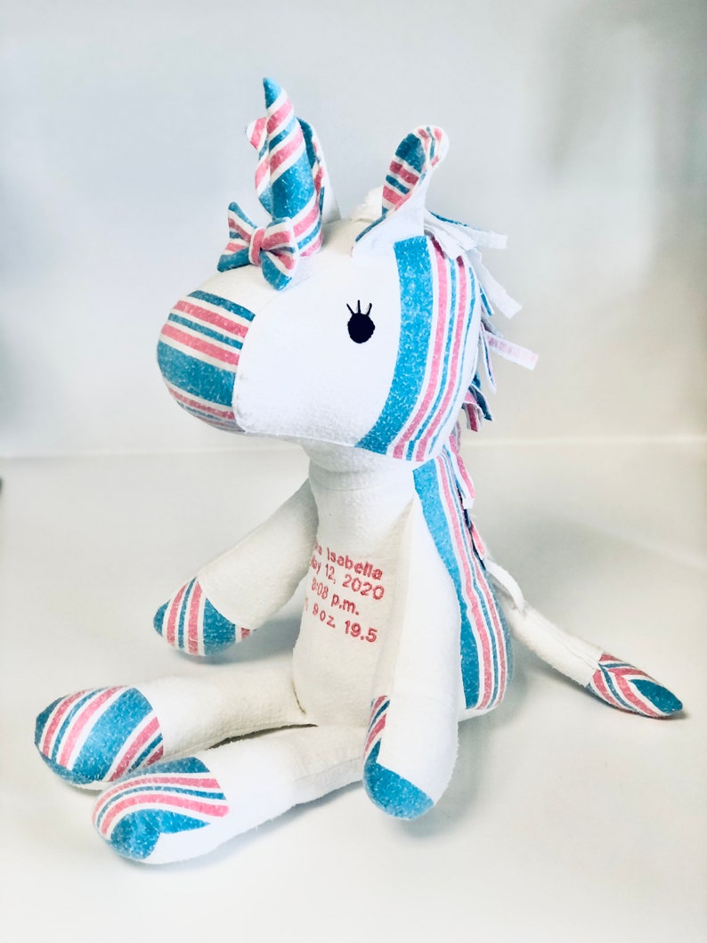 May include: A white unicorn stuffed animal with pink and blue striped fabric. The unicorn has a pink bow on its horn and a black eye. The unicorn is sitting on a white surface. The text "Isabella May 12, 2020 8:08 p.m. 7 oz. 19.5" is printed on the unicorn's body.