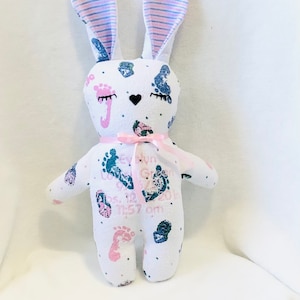 May include: A white bunny stuffed animal with pink and blue striped ears. The bunny is covered in pink and blue footprints and has the text "Evelyn Grace" and "Born 05.12.2018 11:57 am" printed on it.