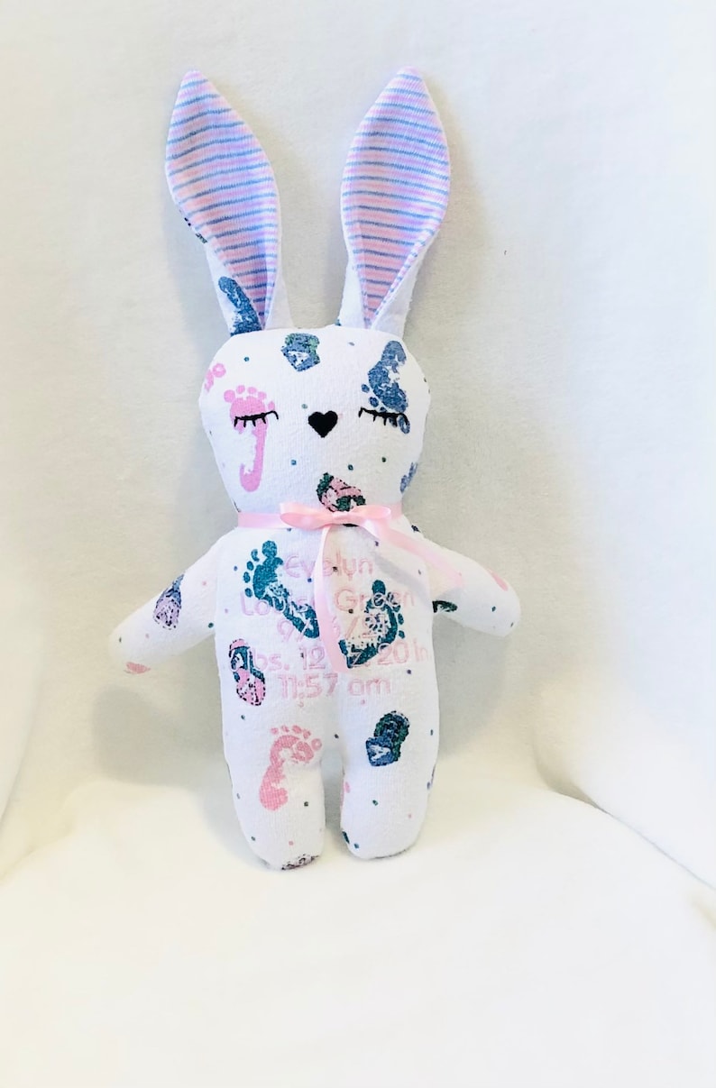 May include: A white bunny stuffed animal with pink and blue striped ears. The bunny is covered in pink and blue footprints and has the text "Evelyn Grace" and "Born 05.12.2018 11:57 am" printed on it.