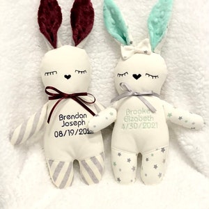 May include: Two plush bunny toys with embroidered names and birthdates. The bunny on the left has burgundy ears and a gray and white striped body. The bunny on the right has light green ears and a white body with green stars.