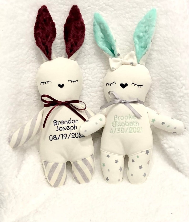 May include: Two plush bunny toys with embroidered names and birthdates. The bunny on the left has burgundy ears and a gray and white striped body. The bunny on the right has light green ears and a white body with green stars.