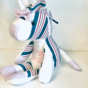 May include: A white unicorn stuffed animal with pink and blue striped fabric. The unicorn has a pink bow and black eyes. The fabric has the text "Little Cate May 5, 2023 19.2 12:03 p.m."