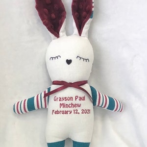 May include: White plush bunny with burgundy ears and blue and white striped arms and legs. The bunny has embroidered eyes, a heart nose, and the text "Grayson Paul Minchew February 12, 2021".