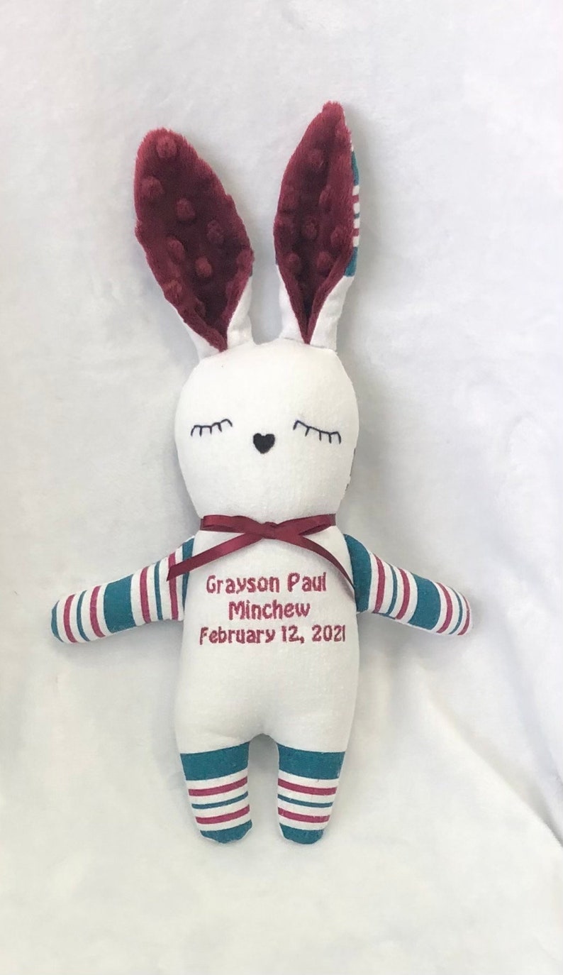 May include: White plush bunny with burgundy ears and blue and white striped arms and legs. The bunny has embroidered eyes, a heart nose, and the text "Grayson Paul Minchew February 12, 2021".