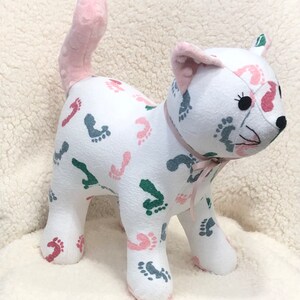 K&ouml;nnte beinhalten: Ein wei&szlig;es Pl&uuml;schkatzenspielzeug mit einem rosa Schwanz. Die Katze ist mit einem Muster aus rosa, gr&uuml;nen und grauen Fu&szlig;abdr&uuml;cken bedeckt. Die Katze hat eine rosa Schleife um den Hals.