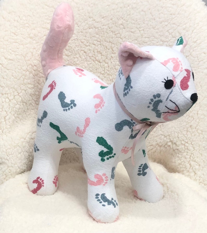 K&ouml;nnte beinhalten: Ein wei&szlig;es Pl&uuml;schkatzenspielzeug mit einem rosa Schwanz. Die Katze ist mit einem Muster aus rosa, gr&uuml;nen und grauen Fu&szlig;abdr&uuml;cken bedeckt. Die Katze hat eine rosa Schleife um den Hals.
