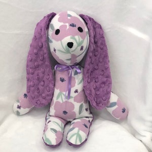 May include: A plush bunny toy with a white body patterned with purple flowers. The bunny has long, textured purple ears and a matching bow. The bunny's face has black eyes and a black nose. The toy is designed for children.