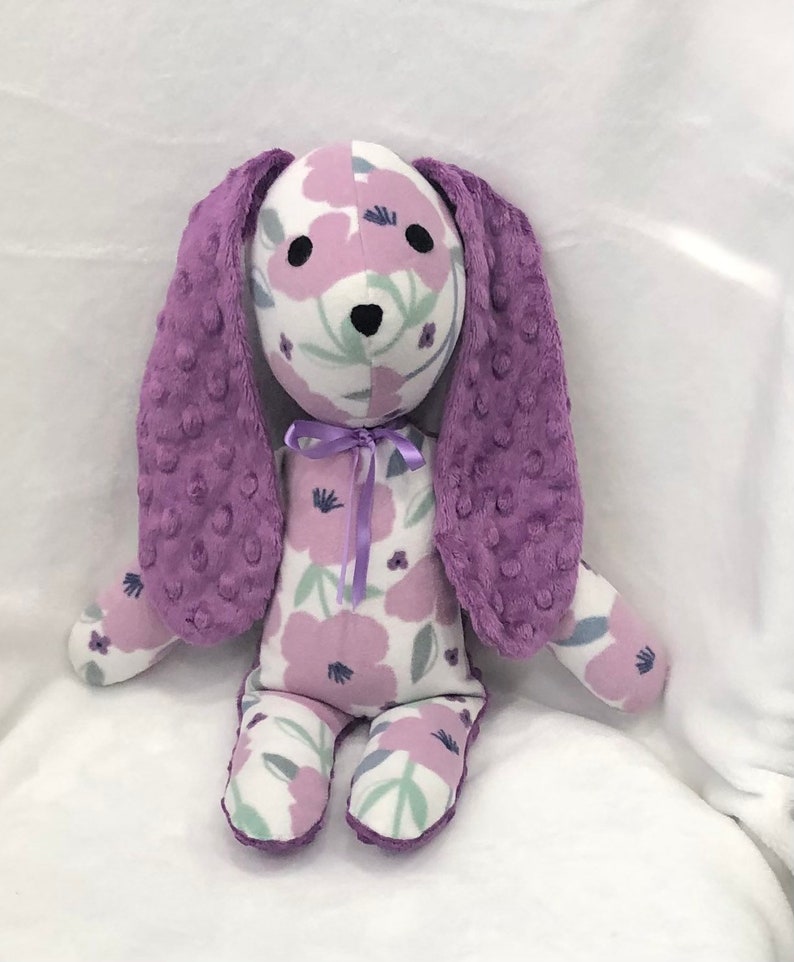 May include: A plush bunny toy with a white body patterned with purple flowers. The bunny has long, textured purple ears and a matching bow. The bunny's face has black eyes and a black nose. The toy is designed for children.