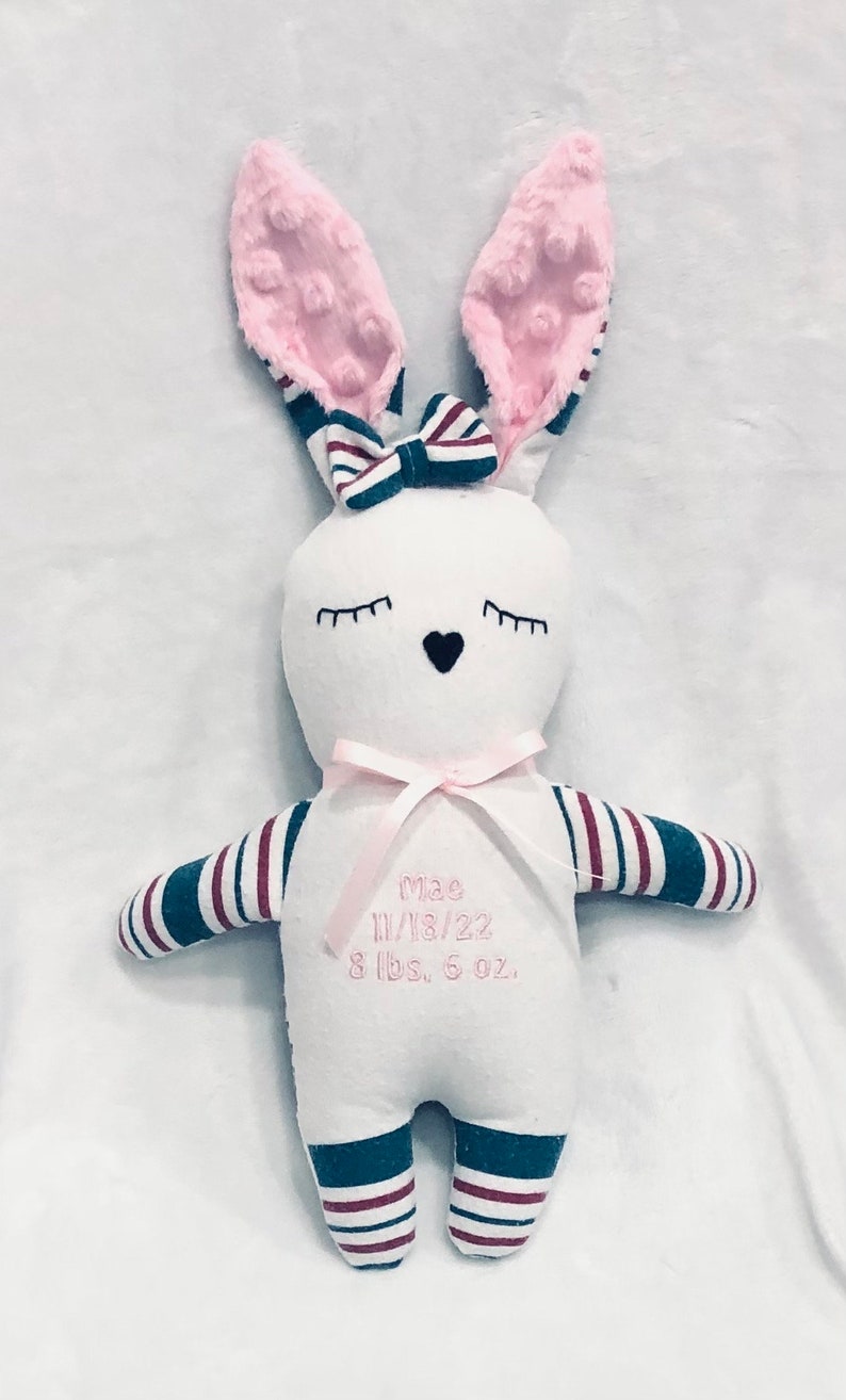 May include: White bunny plush toy with pink ears and a pink bow. The toy has blue and red striped arms and legs. The toy is embroidered with the name "Mae", the date "11/18/22", and the weight "8 lbs. 6 oz."