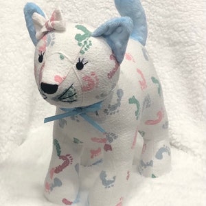 K&ouml;nnte beinhalten: Eine wei&szlig;e Stoffkatze mit einem blauen Schwanz. Die Katze ist mit einem Stoff bedeckt, der ein Muster aus rosa, blauen und gr&uuml;nen Fu&szlig;abdr&uuml;cken hat. Die Katze hat eine blaue Schleife um den Hals und eine rosa Schleife auf dem Kopf.