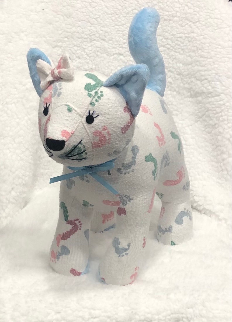 K&ouml;nnte beinhalten: Eine wei&szlig;e Stoffkatze mit einem blauen Schwanz. Die Katze ist mit einem Stoff bedeckt, der ein Muster aus rosa, blauen und gr&uuml;nen Fu&szlig;abdr&uuml;cken hat. Die Katze hat eine blaue Schleife um den Hals und eine rosa Schleife auf dem Kopf.