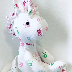 May include: White unicorn stuffed animal with pink, green, and blue handprints and flowers. The unicorn has a white horn and a black eye. The text "lette lurtis 9.09.2021" is printed on the unicorn's body.