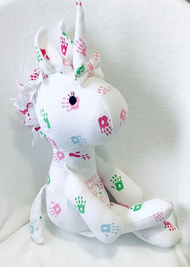 May include: White unicorn stuffed animal with pink, green, and blue handprints and flowers. The unicorn has a white horn and a black eye. The text "lette lurtis 9.09.2021" is printed on the unicorn's body.