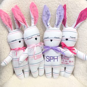 May include: Four white and blue striped bunny plushies with pink, purple, and pink ears. Each bunny has embroidered eyes and a black heart nose. The bunnies have pink, purple, and pink bows around their necks. The bunnies are personalized with the names "Lily", "SPH", and "Jade". The last bunny has the date "6/30/22", weight "5 oz", and length "19 in" embroidered on its chest.