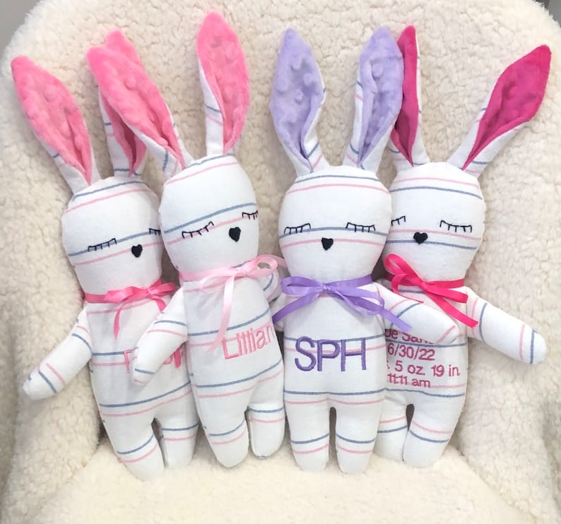 May include: Four white and blue striped bunny plushies with pink, purple, and pink ears. Each bunny has embroidered eyes and a black heart nose. The bunnies have pink, purple, and pink bows around their necks. The bunnies are personalized with the names "Lily", "SPH", and "Jade". The last bunny has the date "6/30/22", weight "5 oz", and length "19 in" embroidered on its chest.