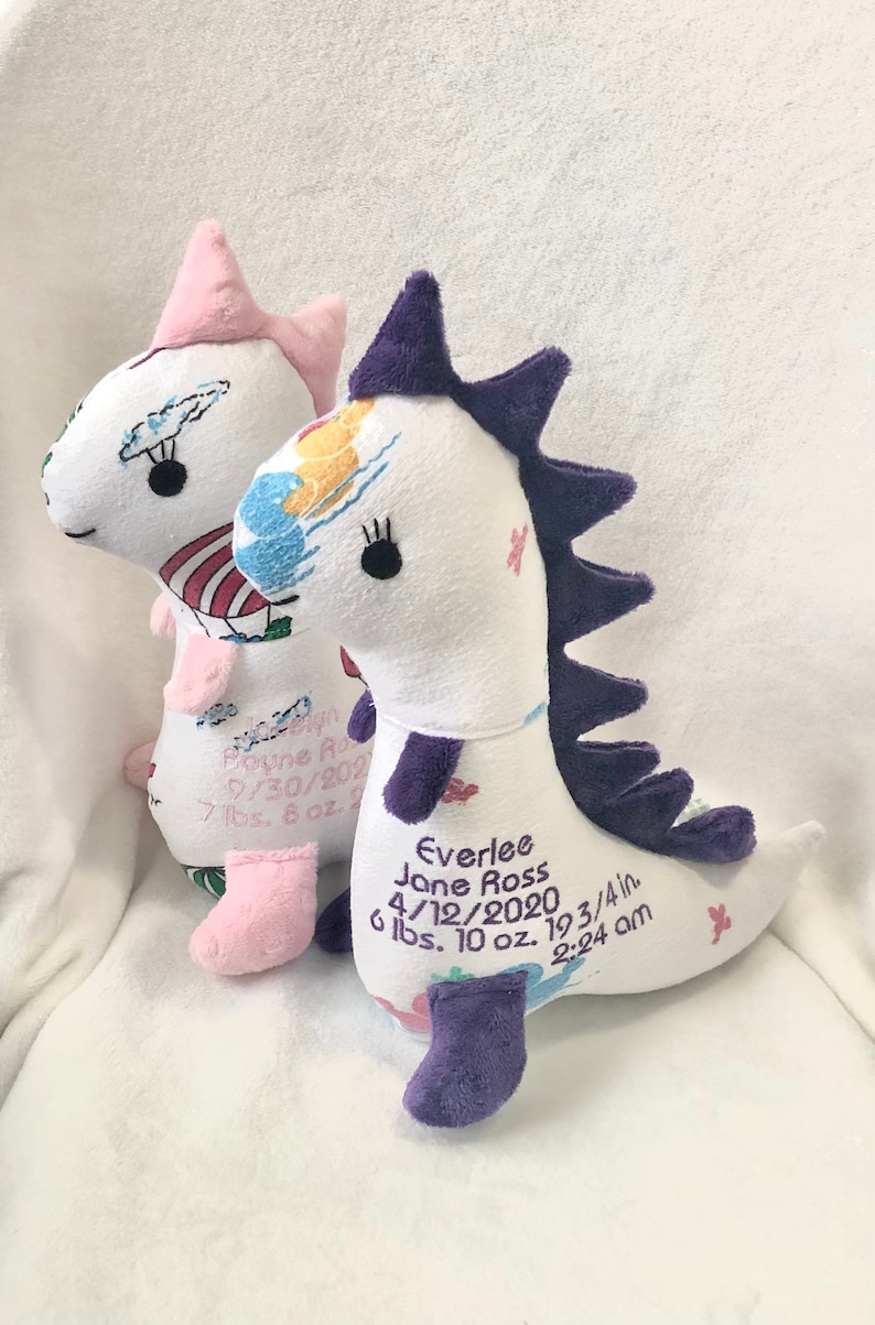 May include: Two white plush dinosaur toys with embroidered details. One has pink accents and the other has purple accents. Each dinosaur has embroidered text with a name, date, weight, and time.