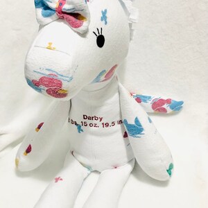 May include: White fabric unicorn stuffed animal with a pink bow, blue and pink floral print, and black eyes. The unicorn is embroidered with the name "Darby" and the text "6 lbs. 15 oz. 19.5 in."