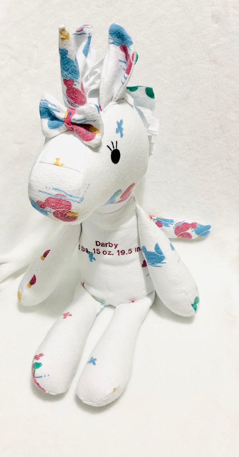 May include: White fabric unicorn stuffed animal with a pink bow, blue and pink floral print, and black eyes. The unicorn is embroidered with the name "Darby" and the text "6 lbs. 15 oz. 19.5 in."