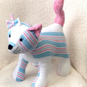K&ouml;nnte beinhalten: Ein Pl&uuml;schtierkatze aus rosa und blau gestreiftem Stoff. Die Katze hat eine rosa Schleife auf dem Kopf und einen rosa Schwanz.