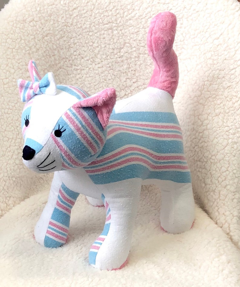 K&ouml;nnte beinhalten: Ein Pl&uuml;schtierkatze aus rosa und blau gestreiftem Stoff. Die Katze hat eine rosa Schleife auf dem Kopf und einen rosa Schwanz.