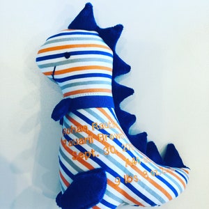 May include: A plush dinosaur toy with blue, orange, and gray stripes. The dinosaur has blue spines and accents. Embroidered text includes a name, date, time, and weight: 9 lbs 8 oz.