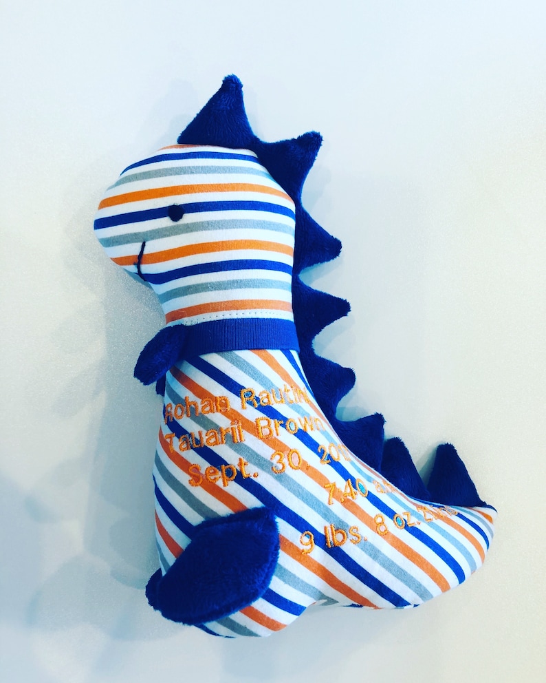 May include: A plush dinosaur toy with blue, orange, and gray stripes. The dinosaur has blue spines and accents. Embroidered text includes a name, date, time, and weight: 9 lbs 8 oz.