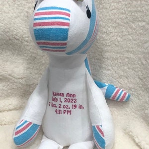 May include: A white unicorn stuffed animal with pink and blue striped fabric. The unicorn has a pink bow with a floral pattern. The unicorn is embroidered with the text "Raven Ann July 1, 2022 7 lbs. 2 oz. 19 in. 4:11 PM".