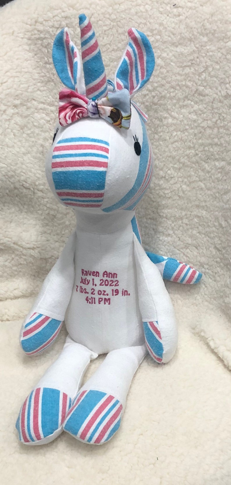 May include: A white unicorn stuffed animal with pink and blue striped fabric. The unicorn has a pink bow with a floral pattern. The unicorn is embroidered with the text "Raven Ann July 1, 2022 7 lbs. 2 oz. 19 in. 4:11 PM".
