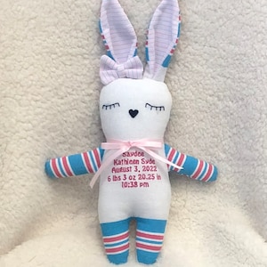 May include: White bunny plush toy with blue and pink striped legs and ears. The bunny has embroidered eyes and a pink bow. The bunny has embroidered text that reads "Saydee Kathleen Syde August 3, 2022 6 lbs 3 oz 20.25 in 10:38 pm".
